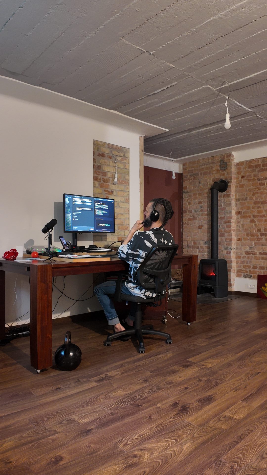Lee at his studio desk, headphones on, wood stove lit, brick walls.
