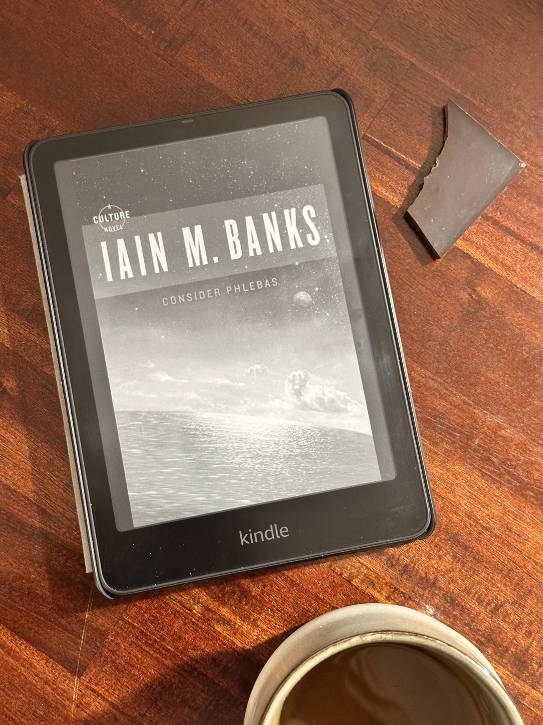 A Kindle on warm wood showing the cover of Iain M. Banks’ Consider Phlebas, a square of dark chocolate and a cup of tea beside it.