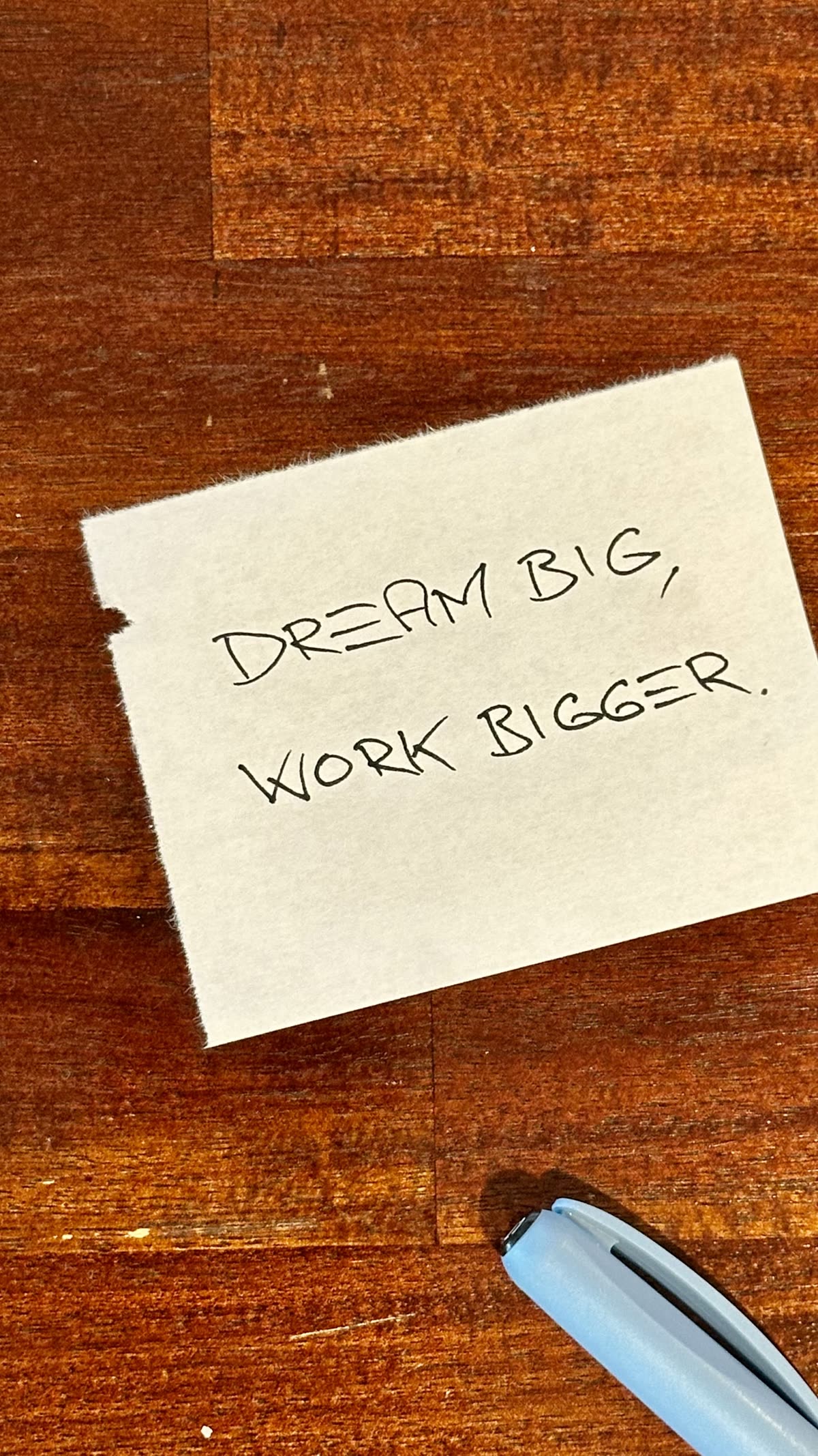 A handwritten note on warm wood, light-blue pen beside it: Dream big, work bigger.