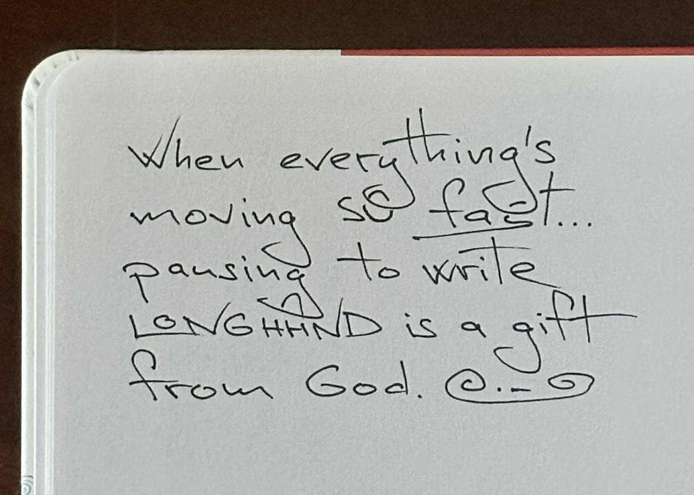 A handwritten journal page on dark wood, white jasmine above: When everything’s moving so fast, pausing to write longhand is a gift from God.