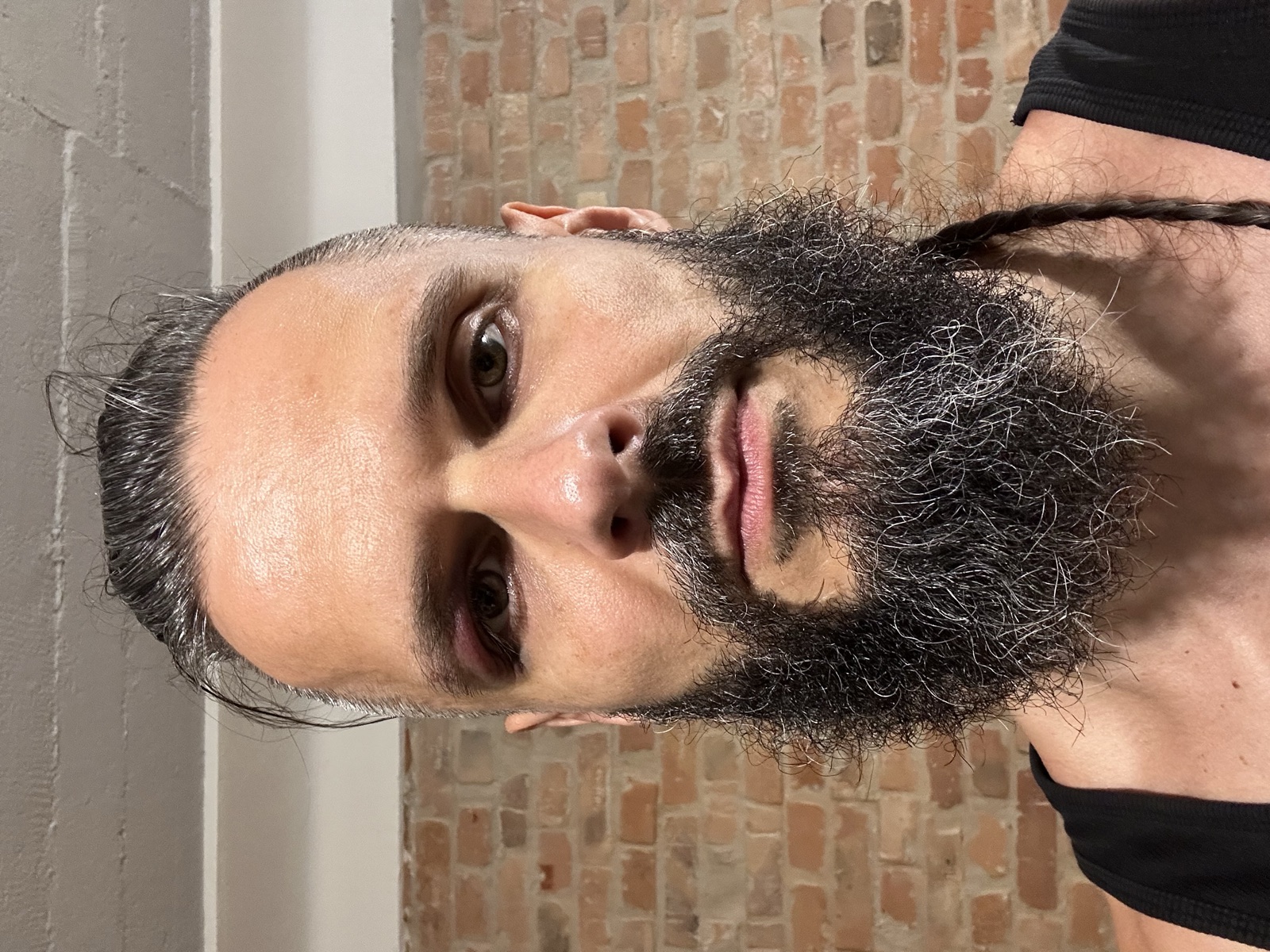 Close-up self-portrait against an exposed brick wall: Lee with a full grey-streaked beard and slicked-back hair, black tank top, level gaze into the lens — a dark purple bruise ringing one eye like smudged eyeshadow, a black eye from impact.