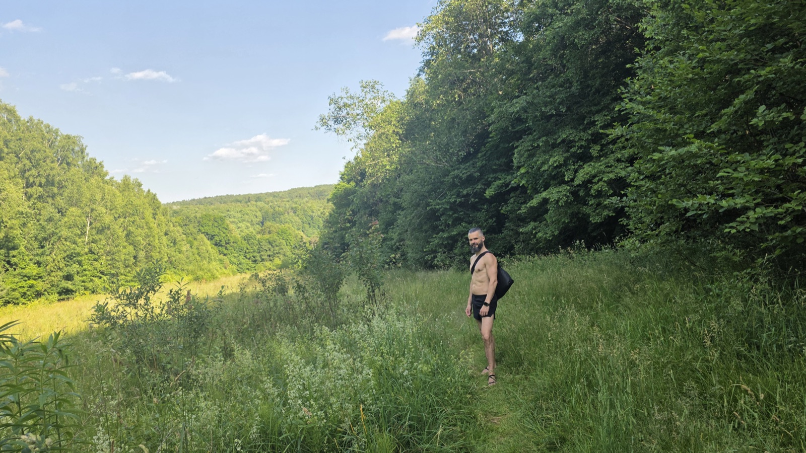 Lee mid-trail in a sunlit Lithuanian meadow, tall green grass and wildflowers underfoot, dense summer forest walls on both sides under a bright blue sky — barefoot-sandals, black shorts, a black sling bag across a bare torso, turning back toward the camera mid-walk.