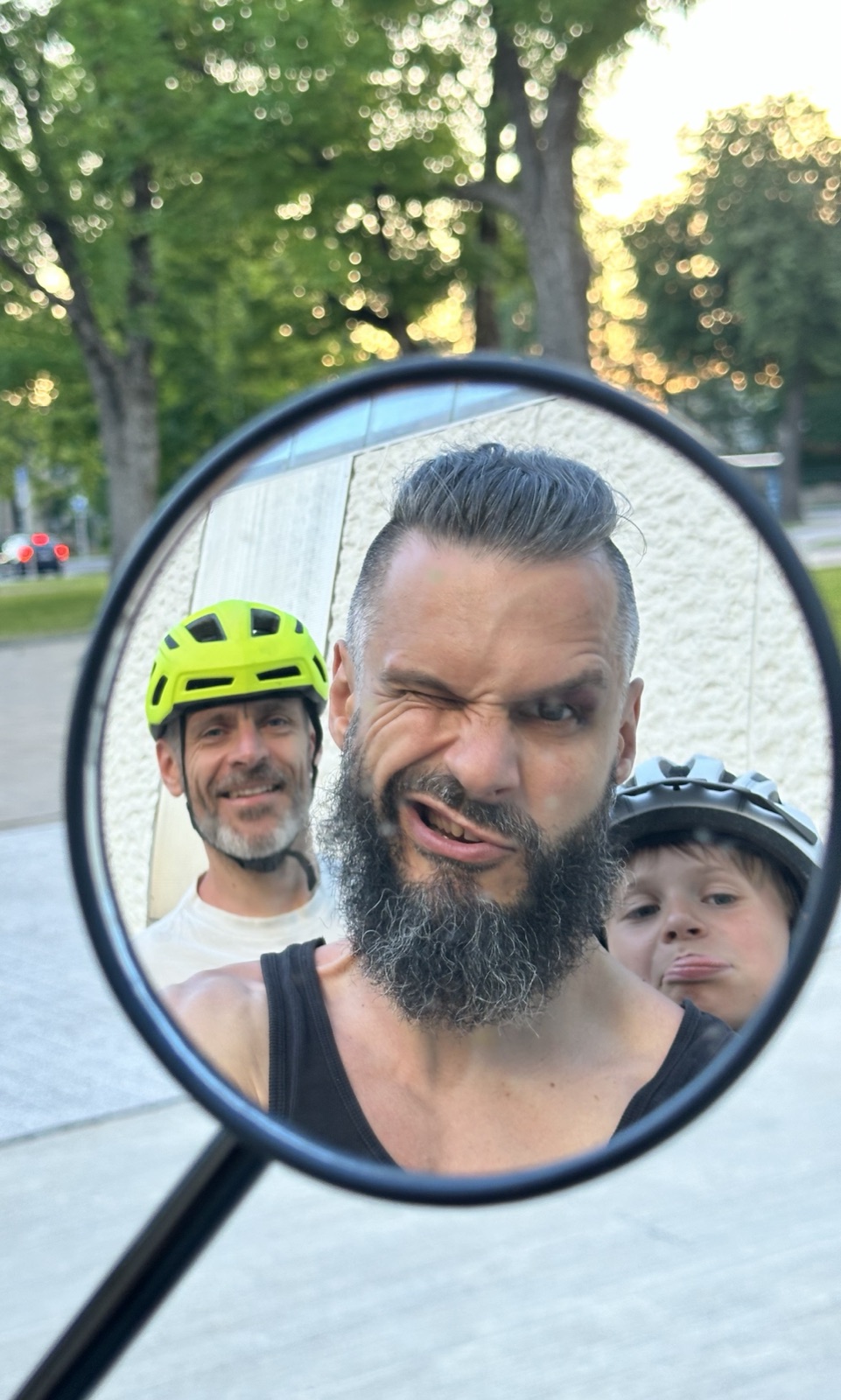 Self-portrait caught in a bicycle's round rearview mirror at golden hour in an urban park: Lee centre-frame in a black tank, big grey beard, hair slicked back, scrunching one eye in a goofy grin; behind him a friend in a lime-green helmet and a kid in a black helmet pulling a face.